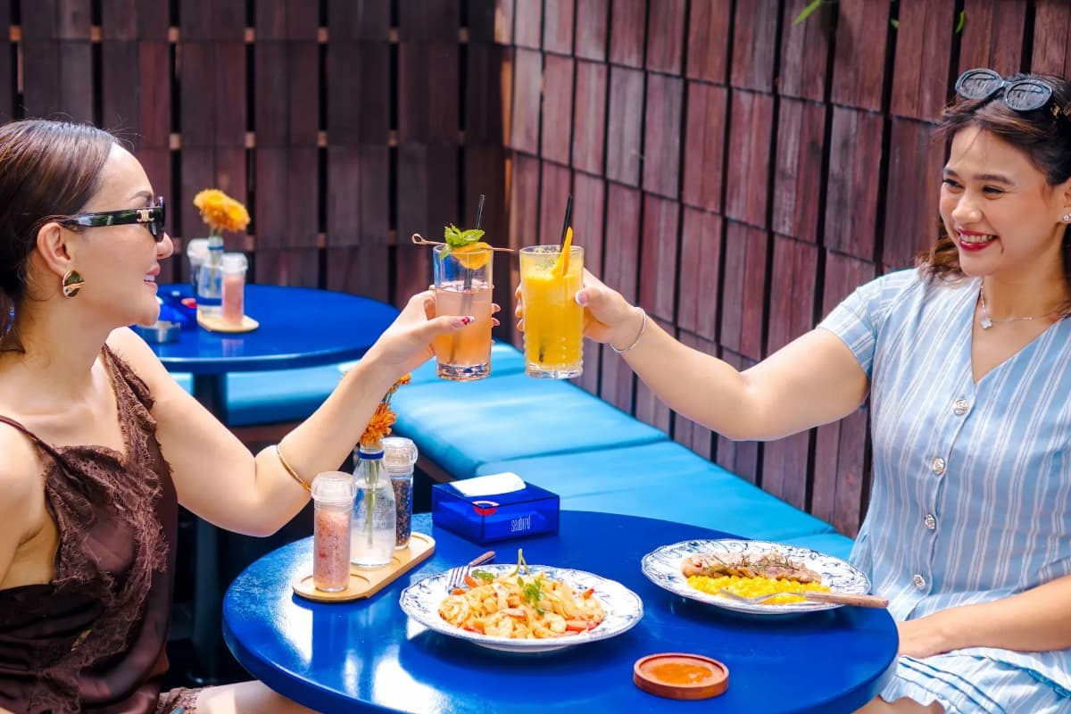 Two women toast drinks at a restaurant table with meals, showing casual dining near Canggu Beach.
