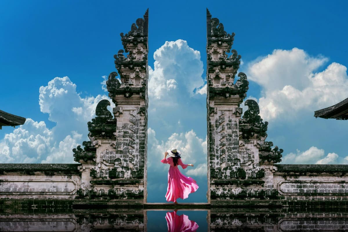 Person standing between Lempuyang temple gates with sky and clouds behind, reflecting on water below.