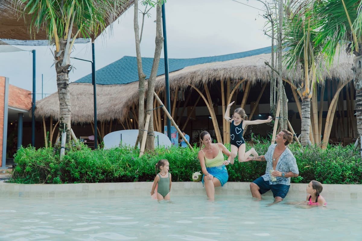 Family with children playing in a shallow resort pool in Bali, surrounded by tropical buildings and palm trees.