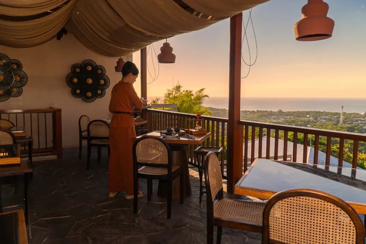Restaurant staff prepares a dining table beside ocean views during sunset at a Bali hillside restaurant.